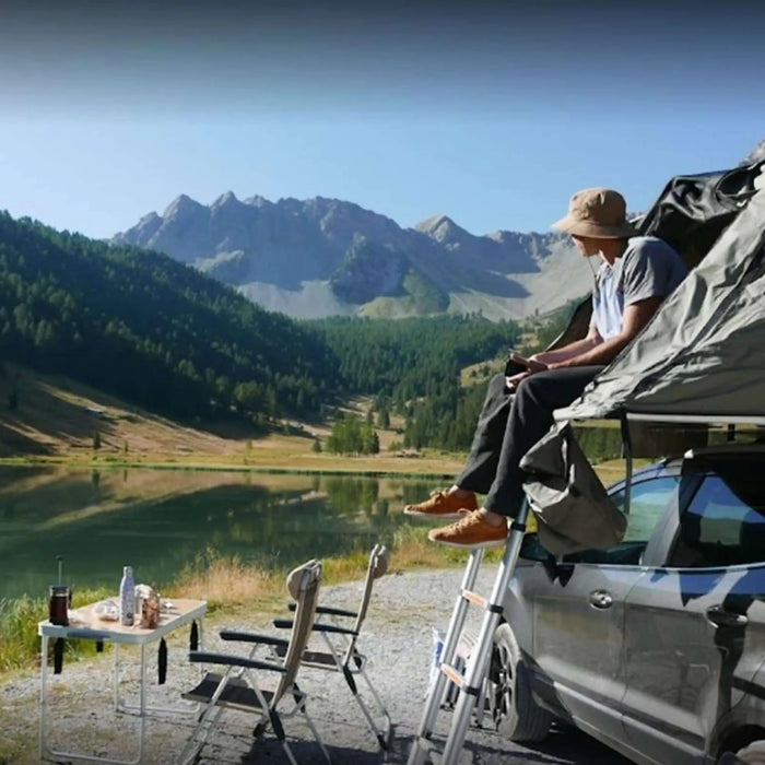 Un homme se relaxant sur le toit d'une tente de voiture près d'un lac pittoresque et des montagnes.