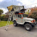 Véhicule 4x4 avec tente de toit rigide pour 2-3 personnes, idéal pour des aventures de camping en plein air, échelle attachée, garé dans une allée.